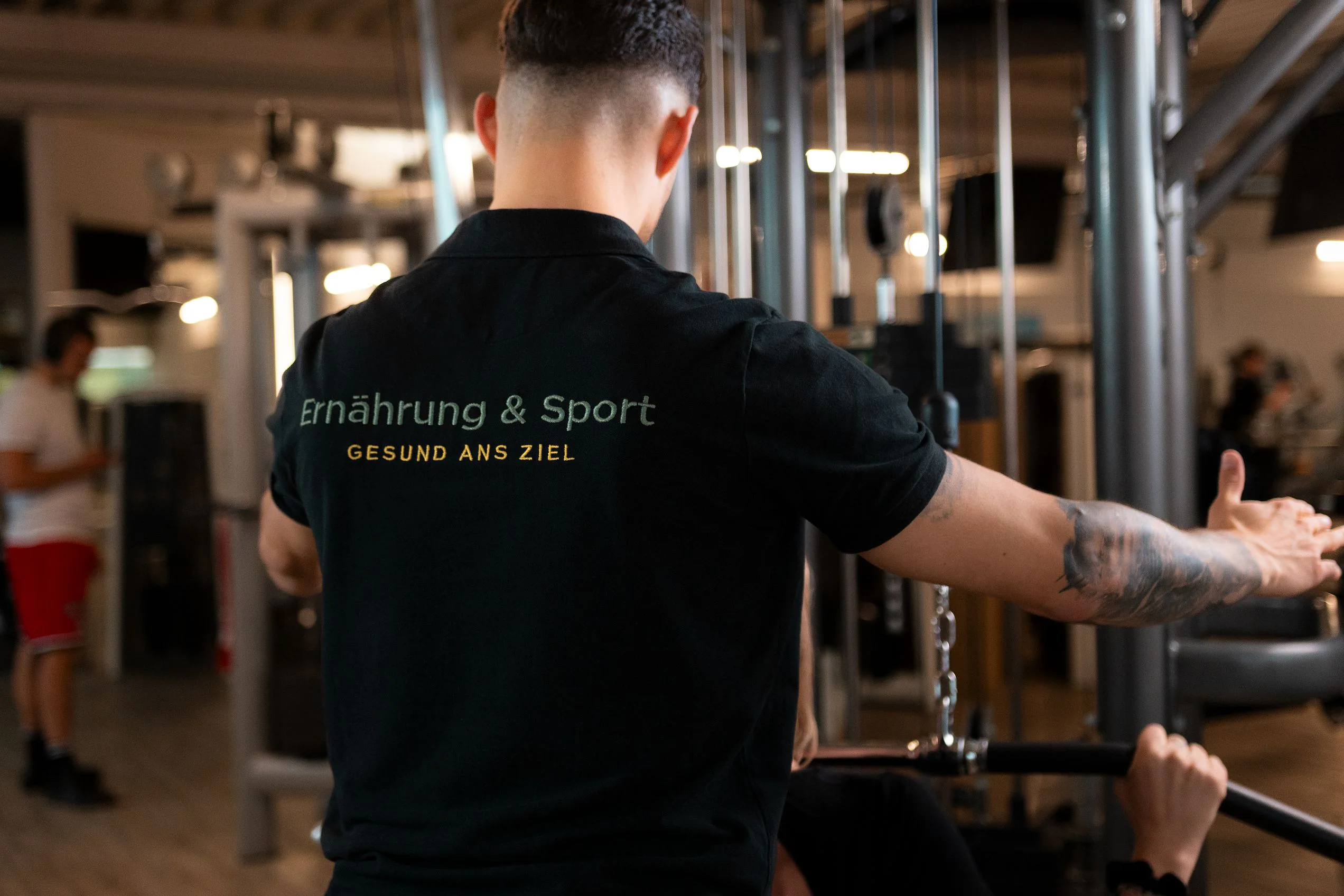 Coach von hinten im Fitnessstudio, auf dem Shirt steht «Ernährung & Sport»